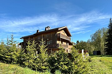A large, multi-story wooden chalet with balconies, surrounded by green trees and grass under a bright blue sky.