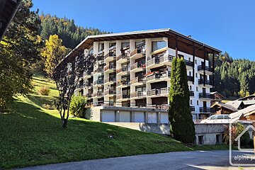An alpine-style apartment building with wooden balconies stands on a grassy slope under a clear blue sky.