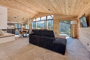 Modern chalet living room featuring light wood paneling, a large black sofa, stone floors, and beautiful mountain views.