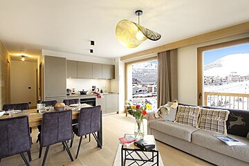 A chic open-plan apartment with dining, kitchen, and living areas. Large windows offer stunning snowy mountain and village views.
