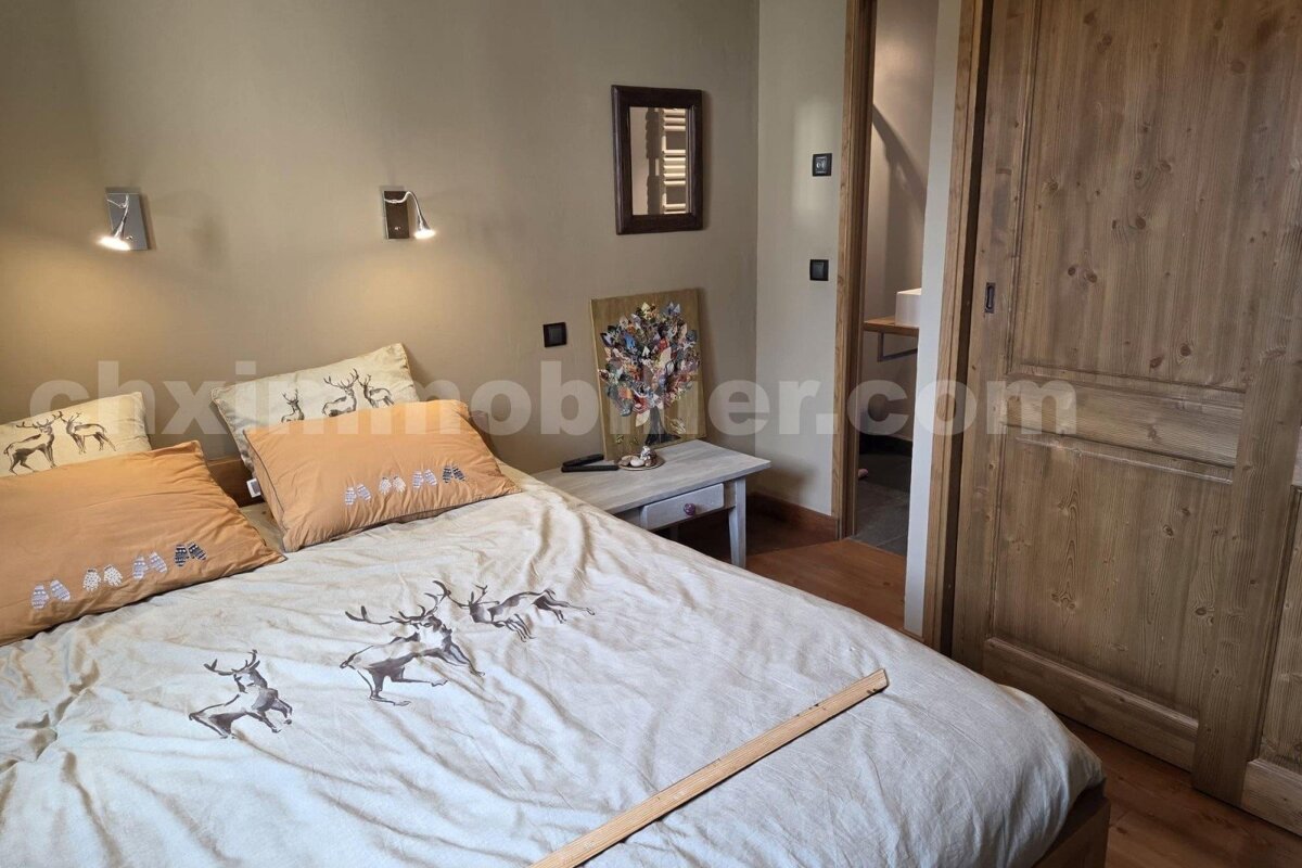A cozy, rustic bedroom with a bed featuring deer-patterned bedding, a nightstand, and warm wooden paneling. A bathroom entrance is visible.