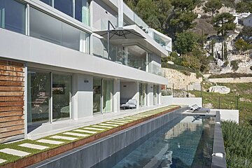 A modern white multi-story villa with glass balconies overlooks an infinity pool and a lush hillside. A patio with stepping stones borders the pool.