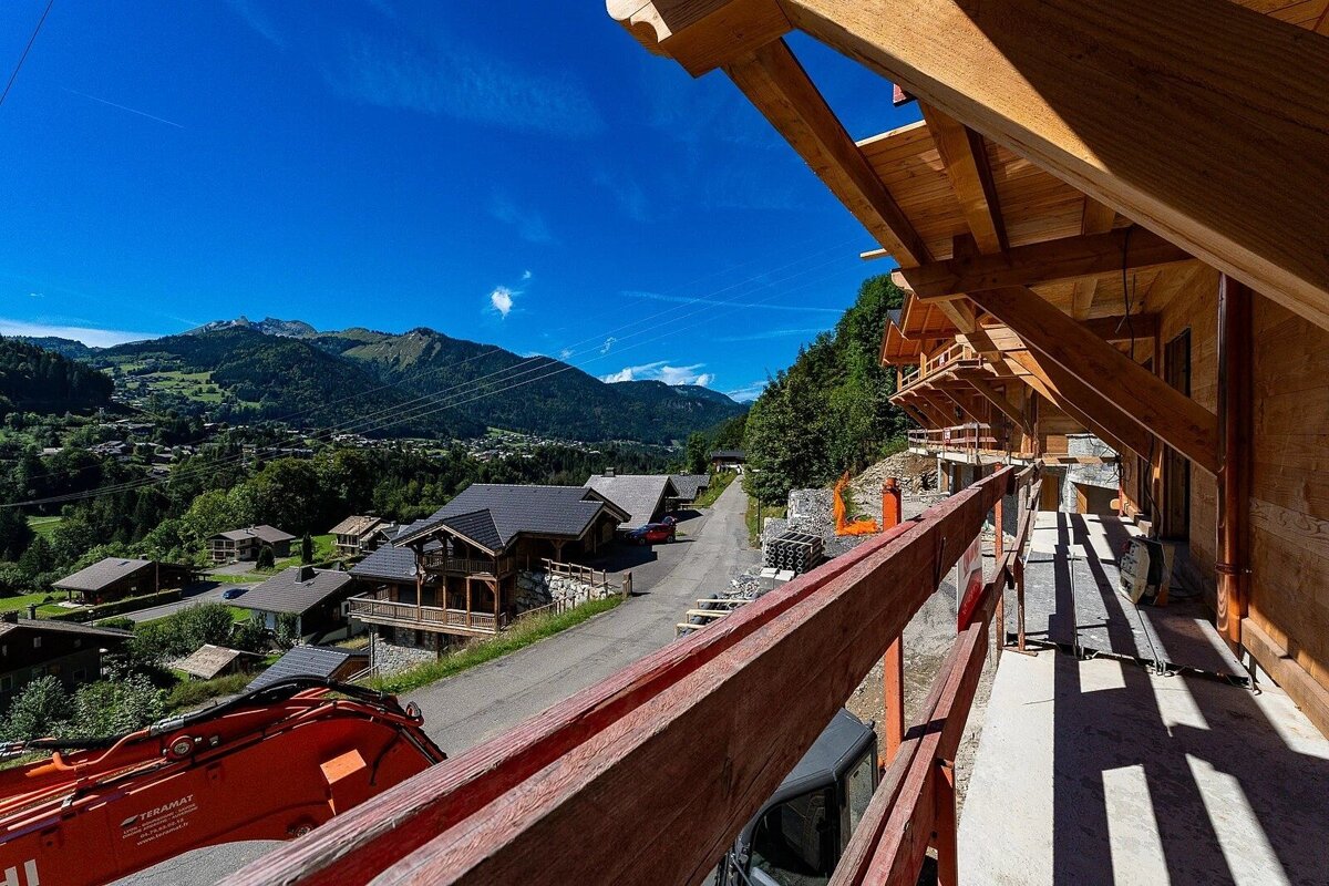A new wooden building under construction with an excavator overlooks a sunny mountain village with chalets and green hills under a clear blue sky.