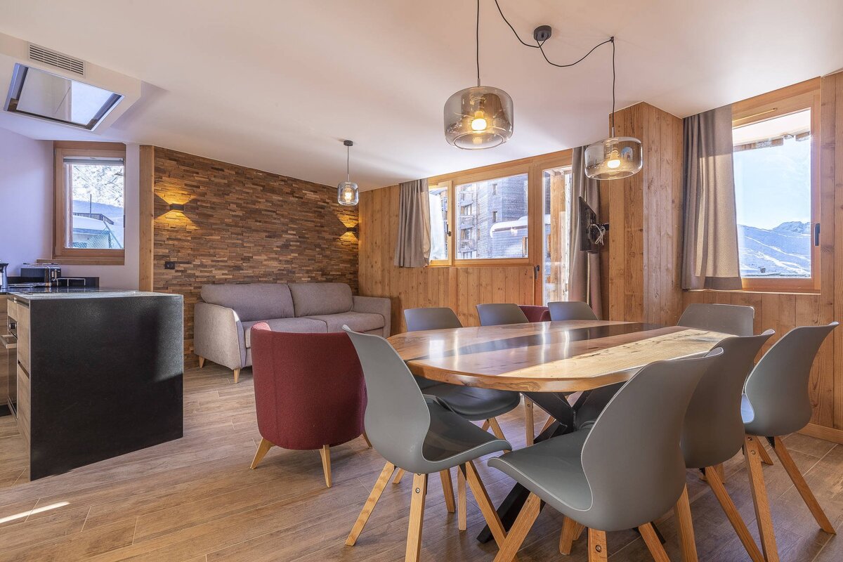 A modern chalet's open-plan dining and living area with a large wooden table, grey chairs, warm lighting, and wood-paneled walls.