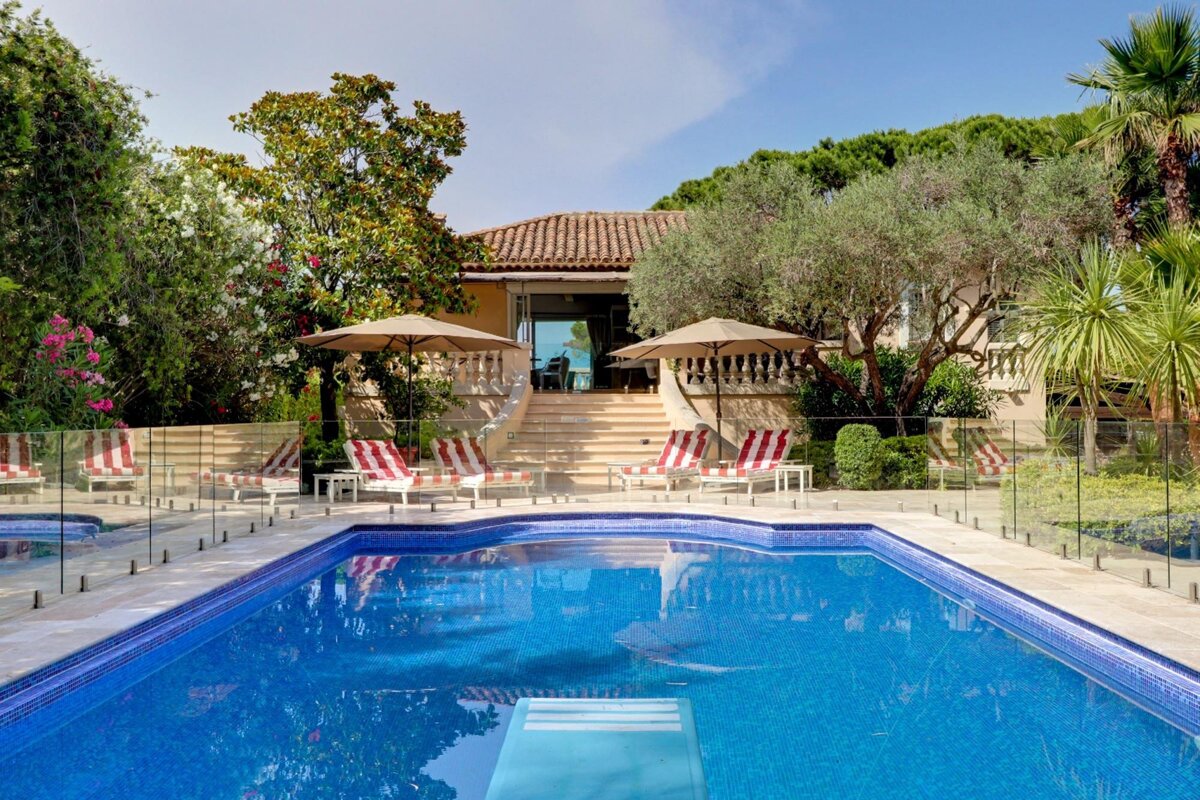 A large swimming pool surrounded by chairs and umbrellas in front of a house