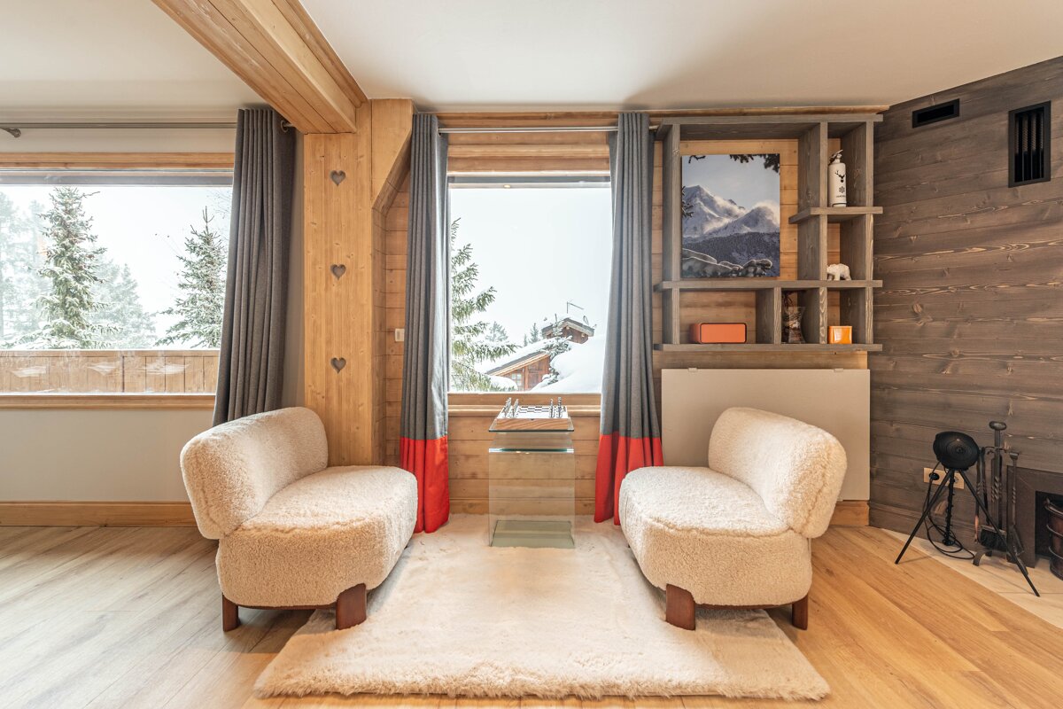 A cozy chalet living room with two white bouclé chairs, wood paneling, a white rug, and large windows revealing a snowy forest outside.