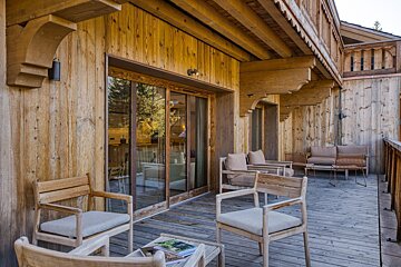 A cozy wooden chalet balcony with outdoor seating and large glass doors, reflecting trees and sunlight.