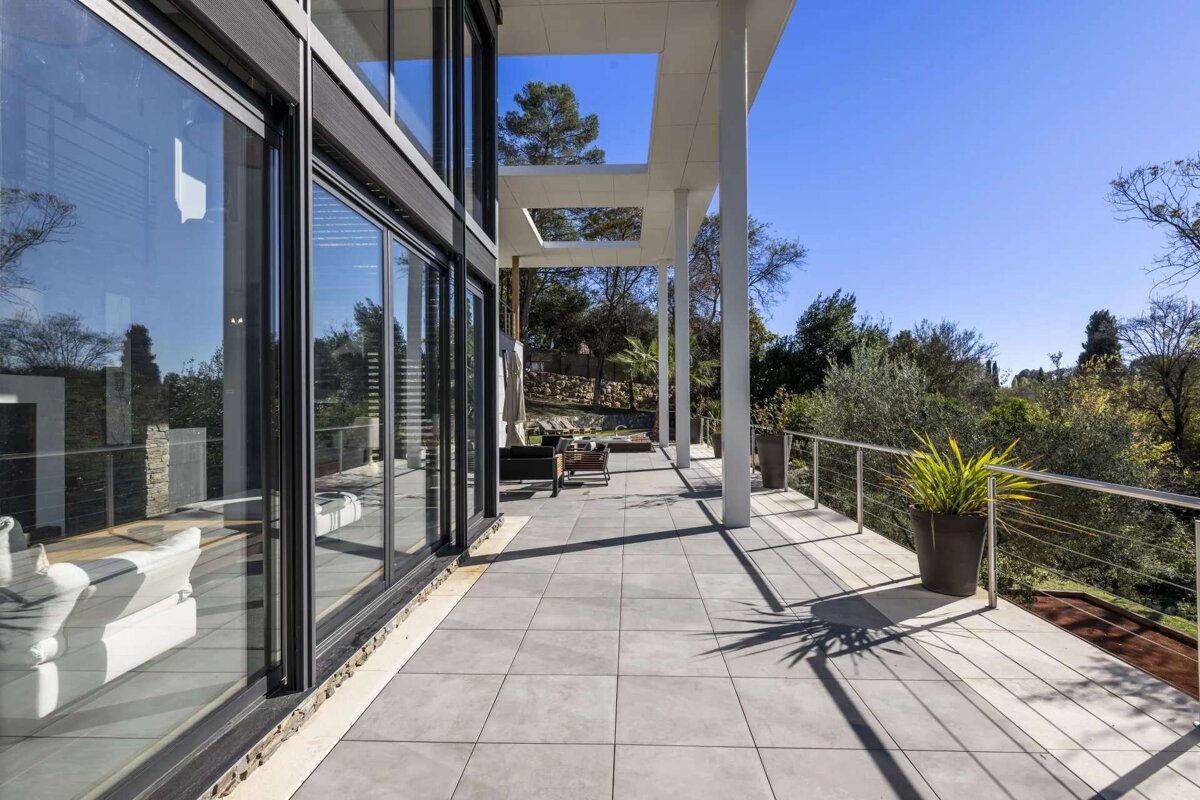 A sleek, modern house with floor-to-ceiling glass doors opens onto a spacious tiled patio featuring white columns and a lush green landscape view.
