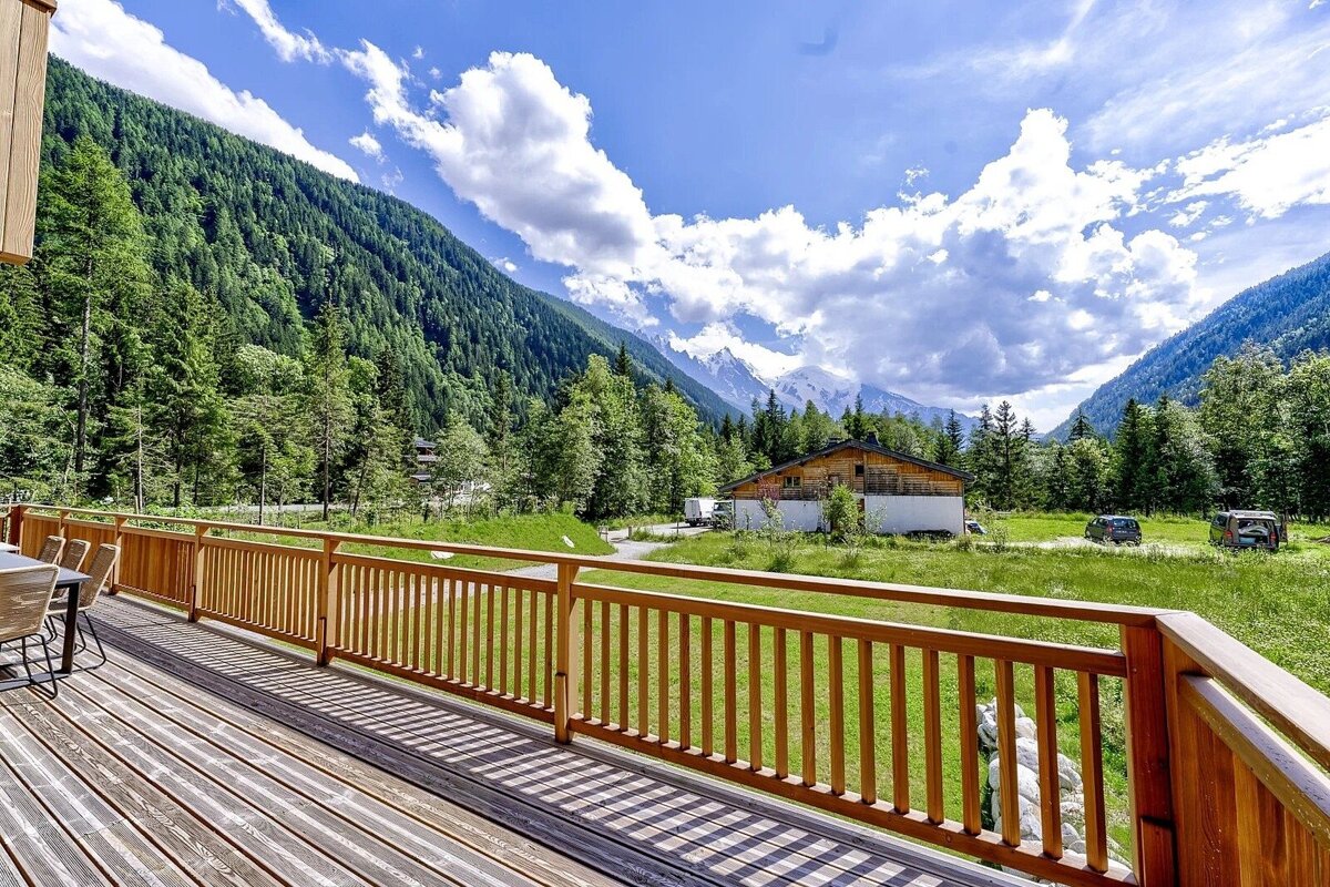 A wooden deck overlooks a green mountain valley with forests, a chalet, and snowy peaks under a blue, cloudy sky.
