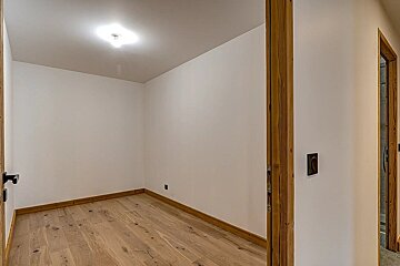 A clean, empty room with white walls, light hardwood floors, and wooden door frames. A ceiling light illuminates the minimalist space.