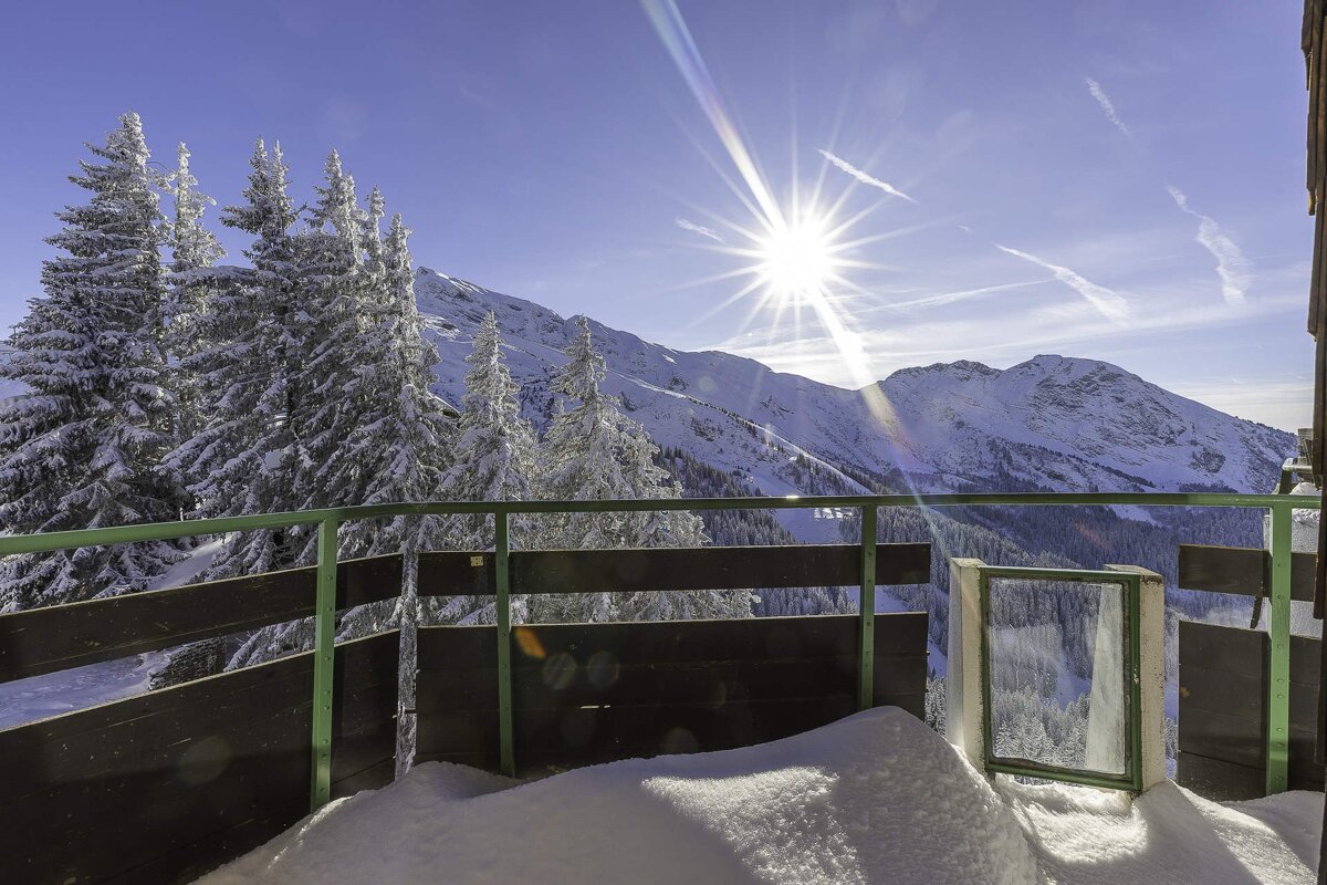 A snow-covered balcony overlooks snow-dusted pine trees and majestic mountains under a bright, sunlit sky.