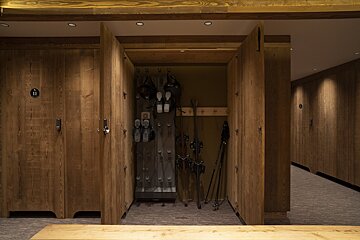 A modern wooden ski locker room with an open cabinet revealing ski boots, skis, and poles. Other locked compartments line the warm, well-lit hallway.