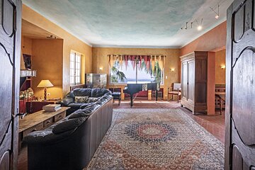 A warm living room with a grand piano by a window, black leather sofa, large patterned rug, and yellow walls, viewed through open doors.