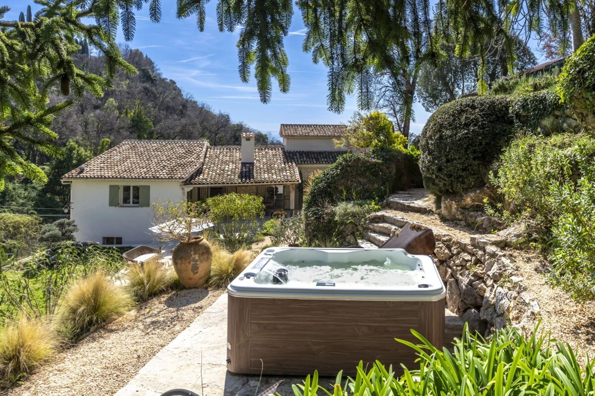 A bubbling hot tub in a lush Mediterranean garden with a white villa, green shutters, and tiled roof, set against a sunny, hilly landscape.