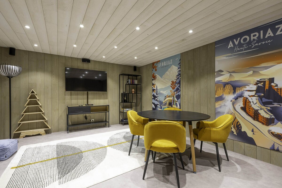 Cozy wood-paneled room featuring a round table with yellow chairs, a wall-mounted TV, and retro Avoriaz ski resort posters.