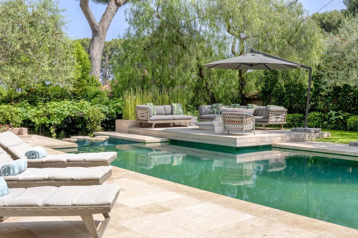 Luxurious backyard pool with sun loungers and a raised patio seating area under a large umbrella, surrounded by lush greenery.