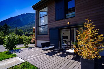 A wooden deck with a couch and a table in front of a house with mountains in the background
