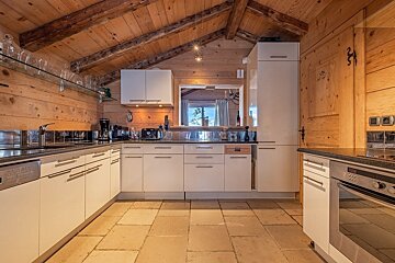 A modern kitchen with white cabinets, dark counters, and stone floor, all surrounded by warm wood walls and ceiling, typical of a rustic chalet.
