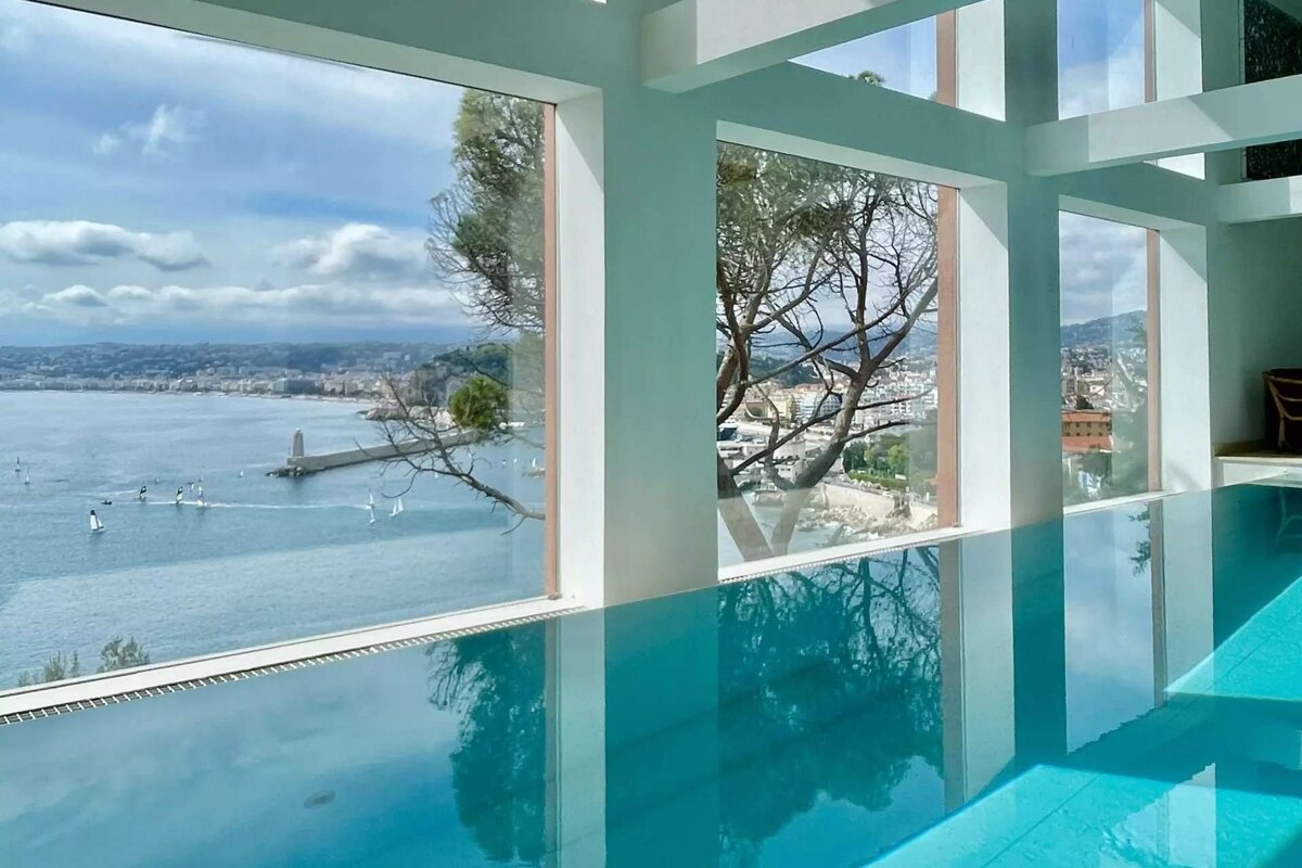 Indoor infinity pool with panoramic sea views of a coastline, lighthouse, and boats through large windows, framed by trees.