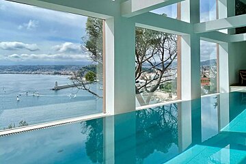 Indoor infinity pool with panoramic sea views of a coastline, lighthouse, and boats through large windows, framed by trees.