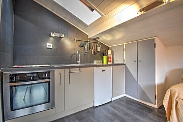 A modern kitchen in an attic space featuring dark tiled walls, grey cabinetry, stainless steel oven, and a slatted wooden ceiling.