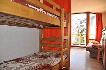 A cozy room features wooden bunk beds, vibrant red curtains, and a large window overlooking a snowy mountain landscape and a balcony.