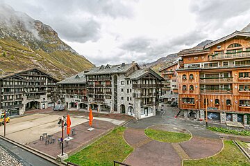 Alpine village square featuring traditional stone and wooden chalets under a cloudy sky with mountain backdrops.