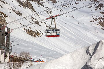 A gondola hangs above a snowy mountain slope, with a building and people below.