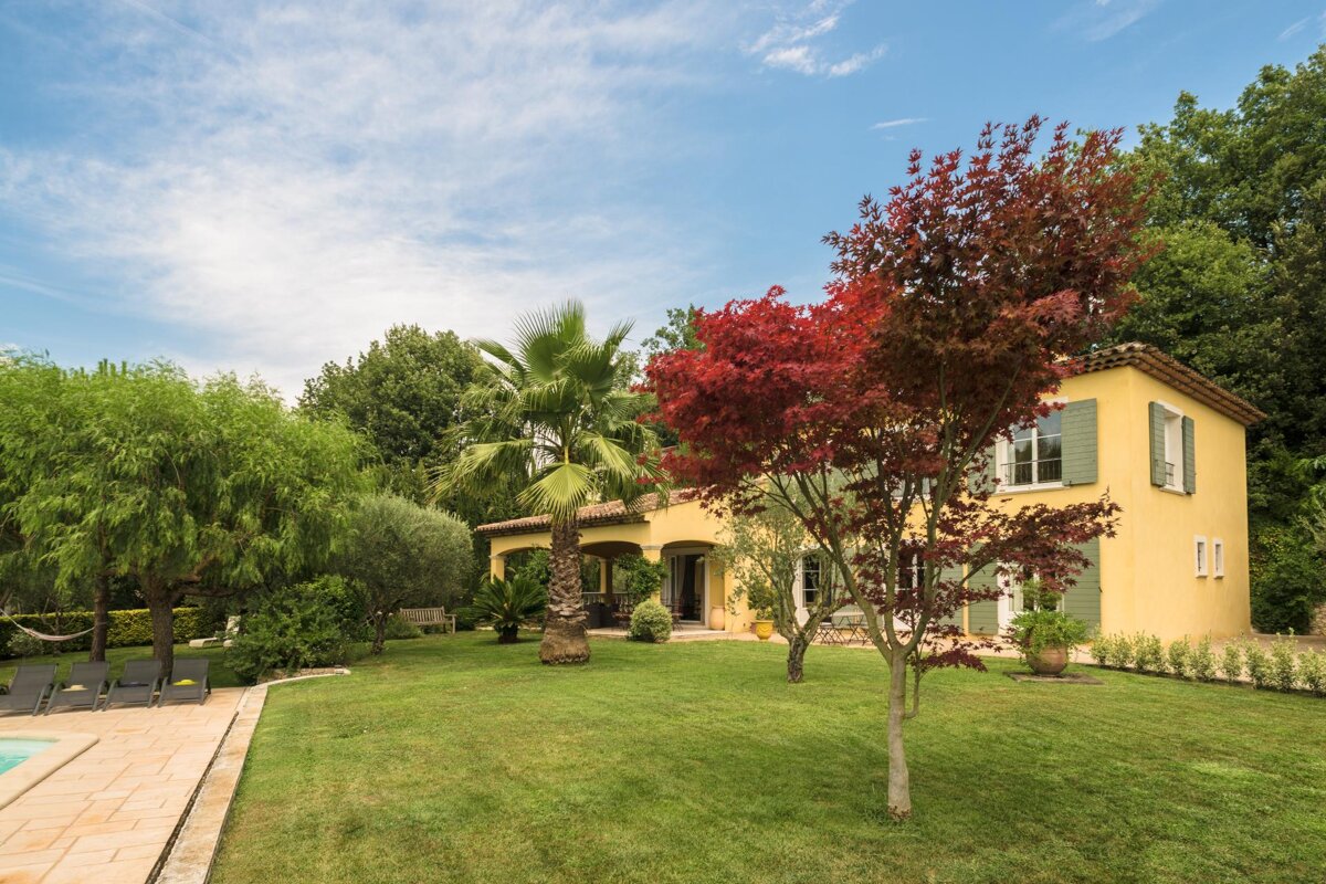 A yellow house with green shutters sits in the middle of a lush green yard