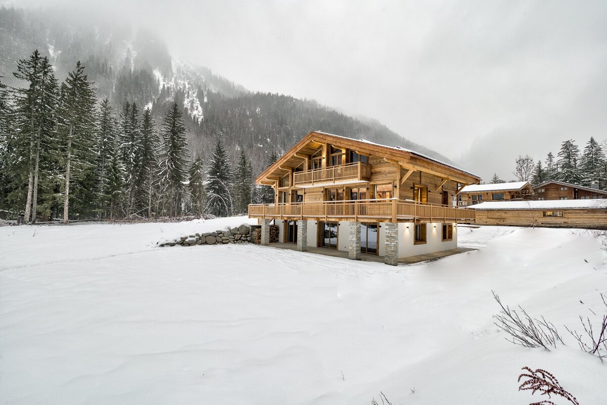 A large, modern wooden chalet is nestled in a snow-covered valley, with pine forests and misty mountains in the background on an overcast day.
