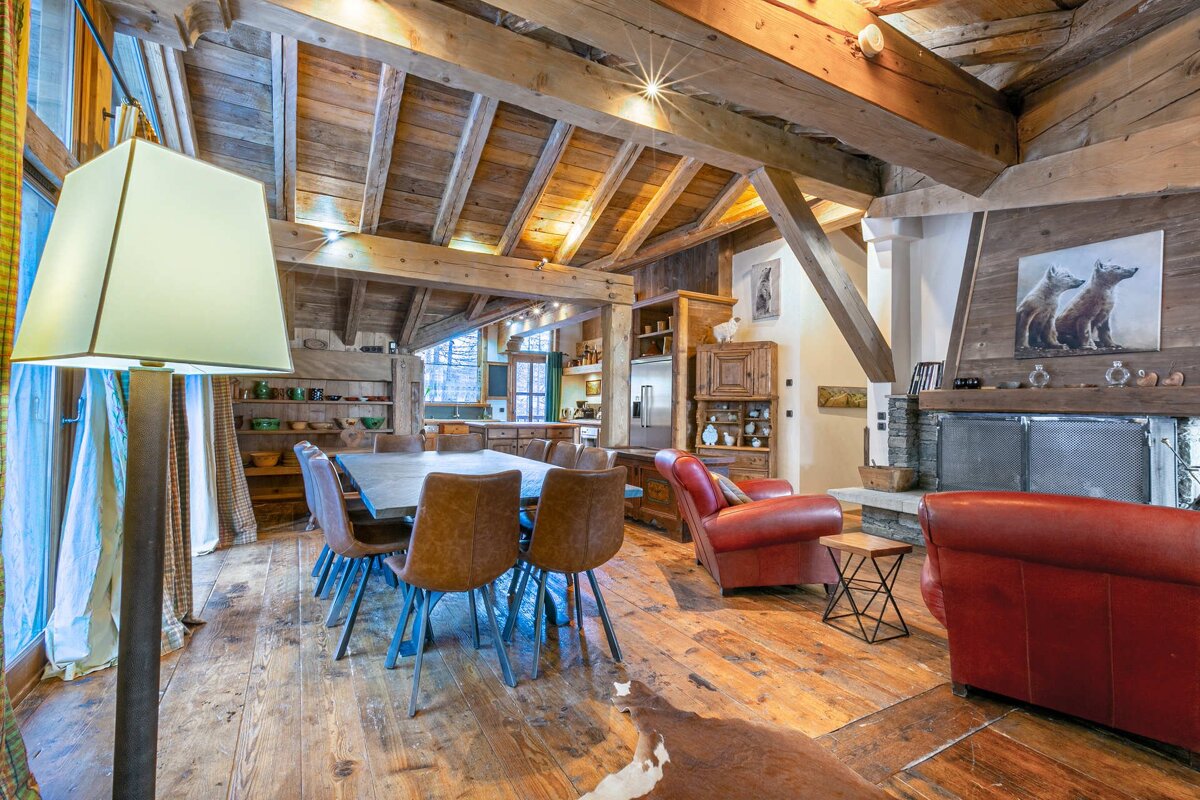 A cozy, rustic wooden interior featuring exposed beams, a dining table, red armchairs, a stone fireplace, and a kitchen.