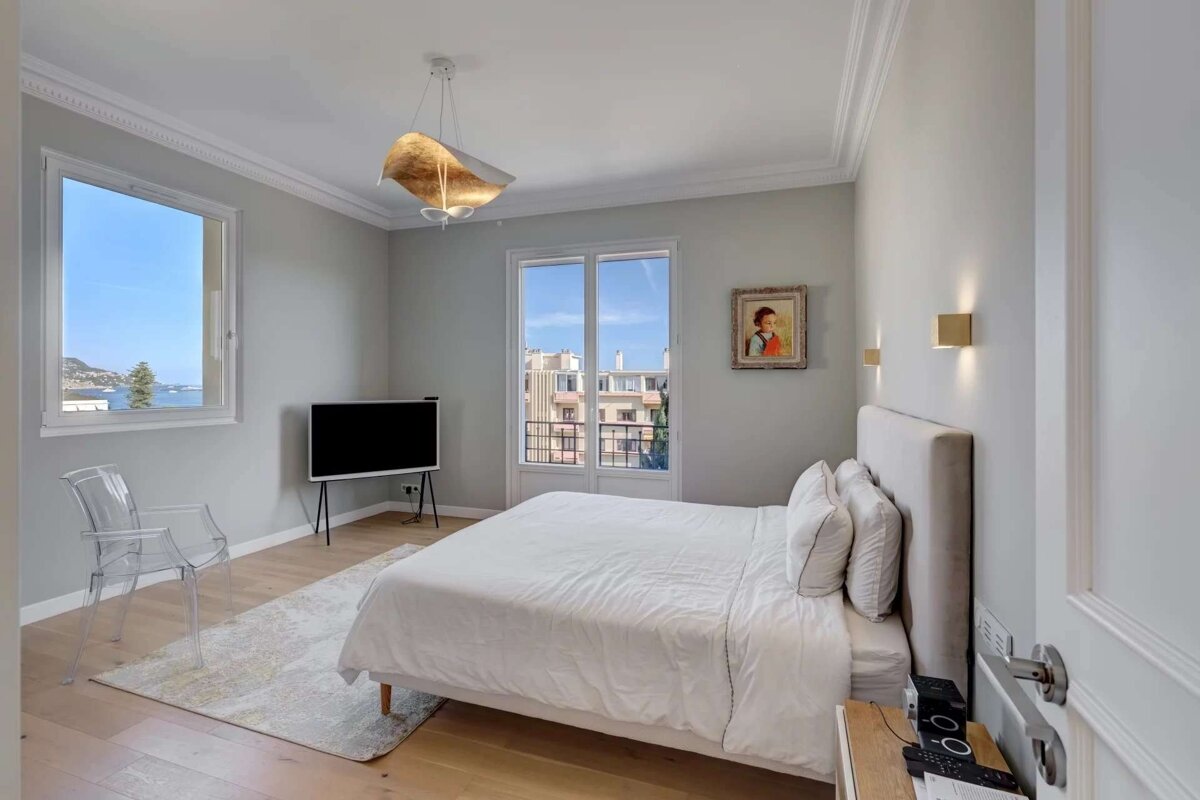 Bright, modern bedroom with large windows offering sea and city views. Features a white bed, TV, clear chair, and light wood flooring.