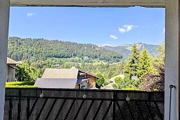 Balcony view looking out over lush green forested mountains and alpine chalets under a bright blue sky.
