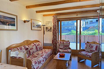 A cozy, rustic living room features wooden furniture with patterned cushions, exposed ceiling beams, and a large sliding glass door leading to a balcony.