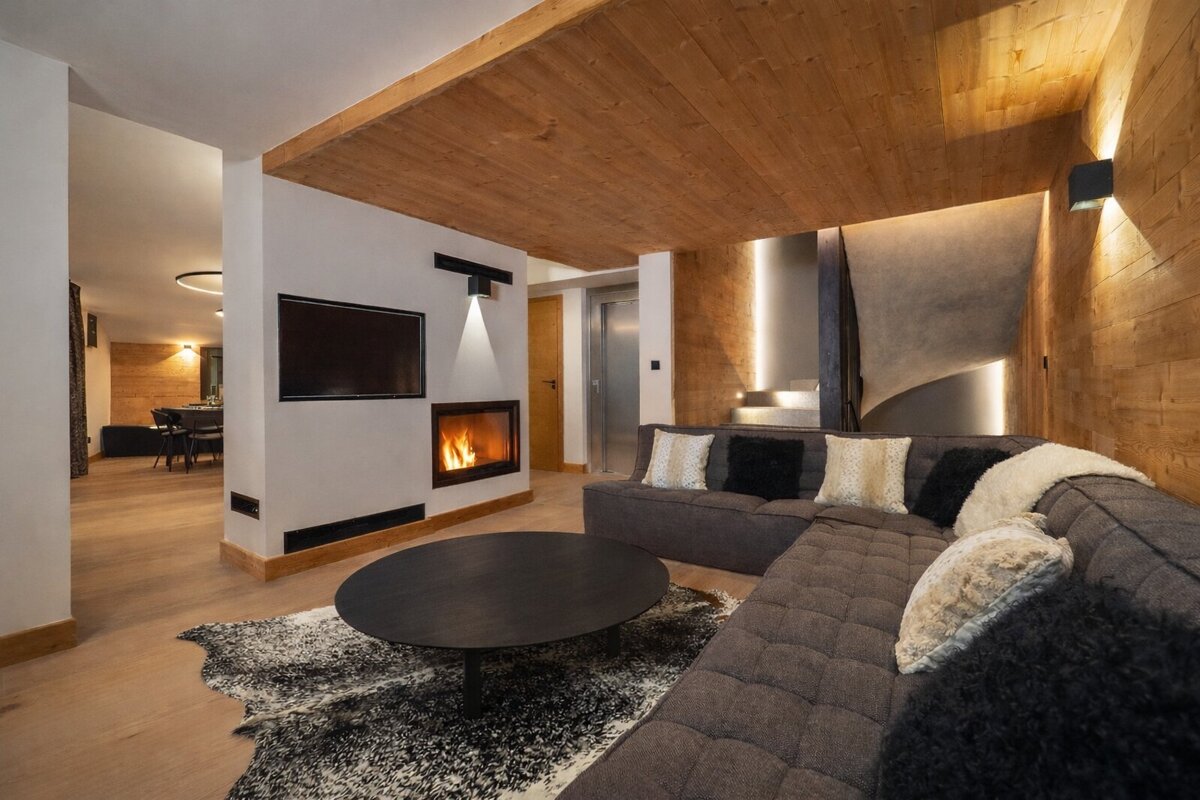 Cozy, modern living room featuring a fireplace, TV, L-shaped sofa, and natural wood accents on ceiling/walls. A dining area and stairs are visible.