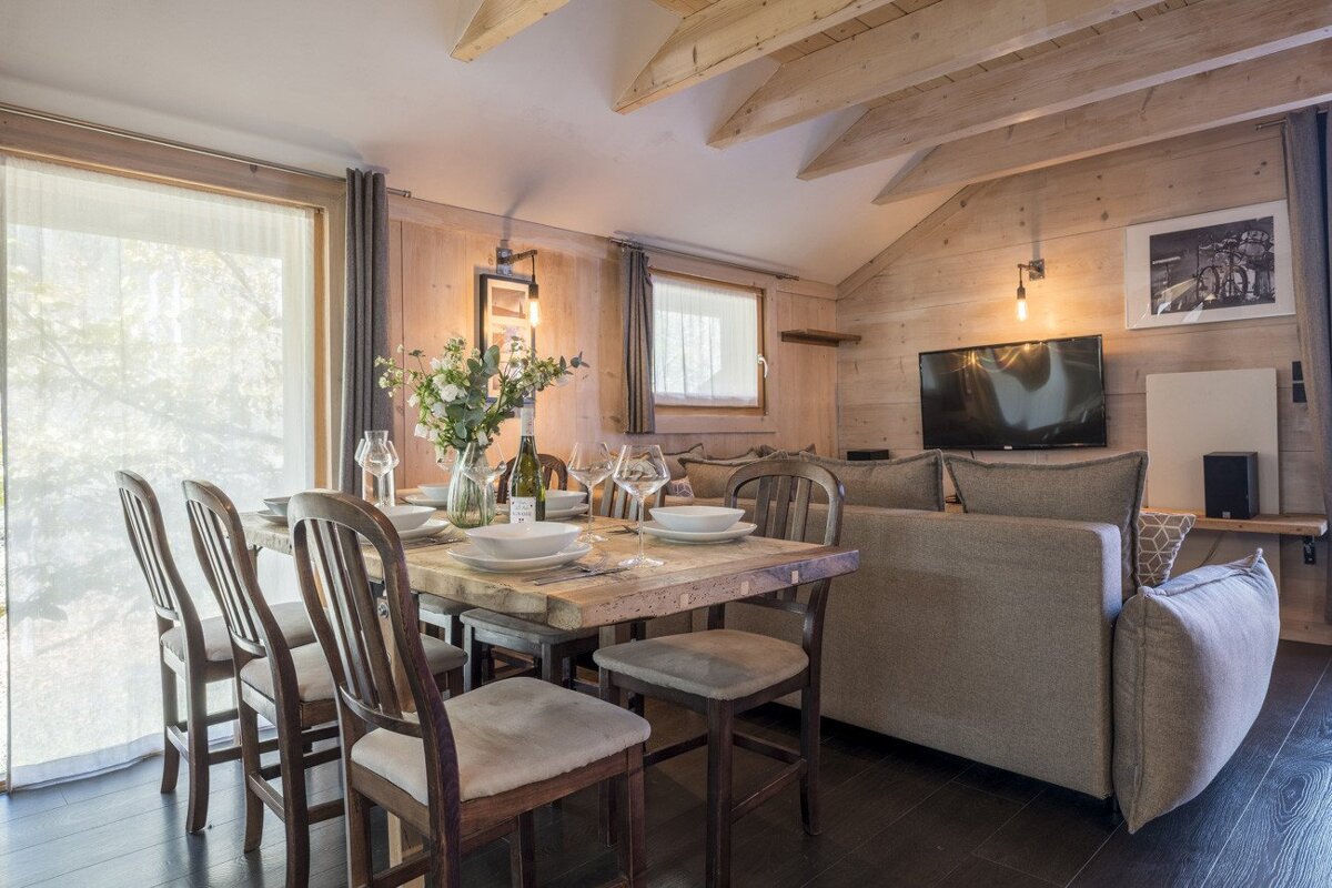 A warm, modern chalet interior with a set dining table, a cozy living area with a TV, and rustic wood paneling throughout.