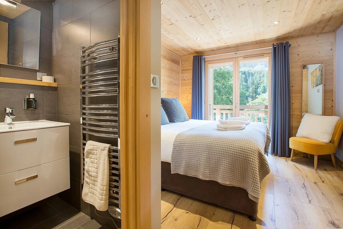 A modern, wood-paneled bedroom with a bed, balcony forest view, and a yellow chair. Adjacent, a sleek bathroom with a vanity and heated towel rack.