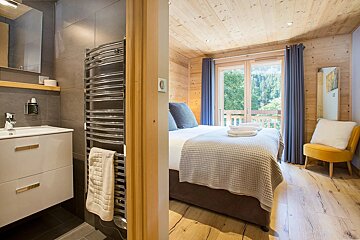 A modern, wood-paneled bedroom with a bed, balcony forest view, and a yellow chair. Adjacent, a sleek bathroom with a vanity and heated towel rack.