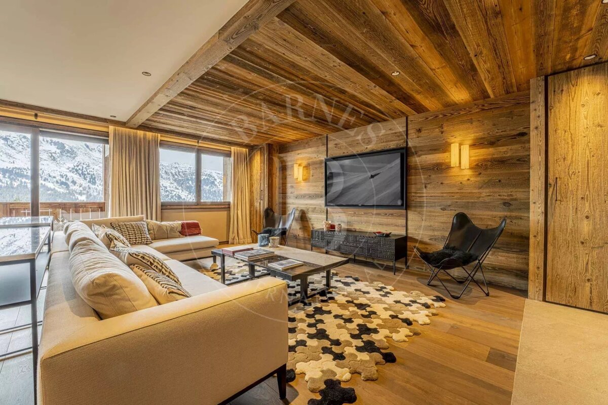 A cozy, modern chalet living room with wood-paneled walls/ceiling, a large sofa, TV, unique rug, and big windows showcasing snowy mountain views.