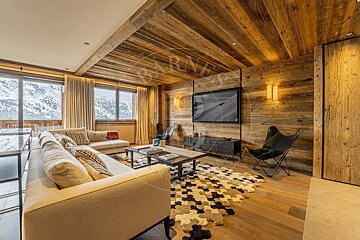 A cozy, modern chalet living room with wood-paneled walls/ceiling, a large sofa, TV, unique rug, and big windows showcasing snowy mountain views.
