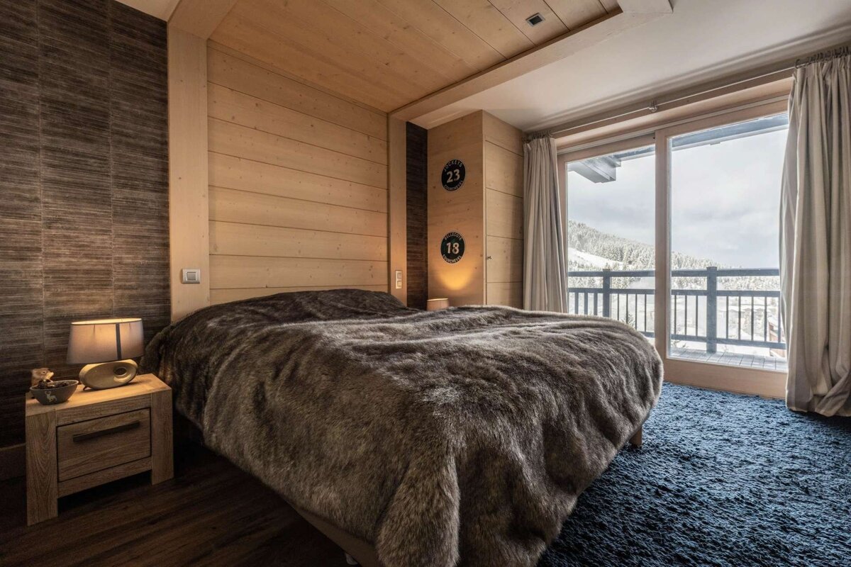 A cozy chalet bedroom with wood paneling, a plush fur blanket on the bed, and a large window revealing a snowy mountain view.