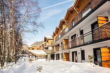 A modern white and wood building with balconies stands in deep snow next to bare trees, under a clear blue sky. It's a sunny winter day at a mountain resort.