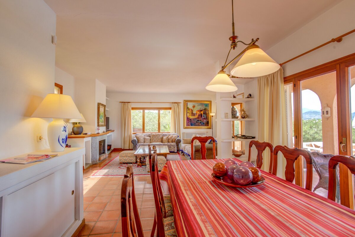 Bright, open-plan living and dining room with terracotta floors, warm wooden furniture, and large windows opening to an outdoor view.