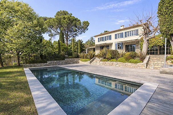 A two-story villa with blue shutters overlooks a large swimming pool surrounded by lush green trees and a wooden deck.