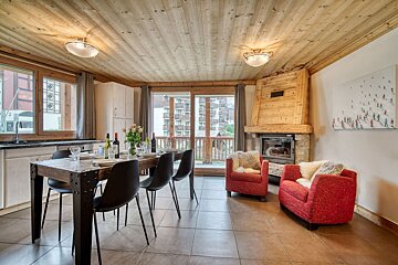 Cozy chalet living/dining room with wooden ceiling, set dining table, red armchairs by a stone fireplace, and a large skier painting.