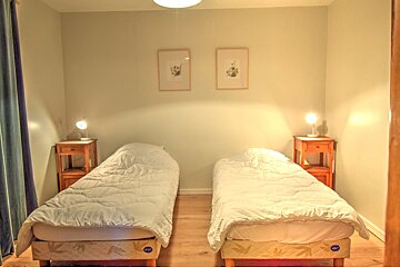 A cozy bedroom features two single beds with white duvets, wooden nightstands with lamps, light green walls, and two framed pictures.
