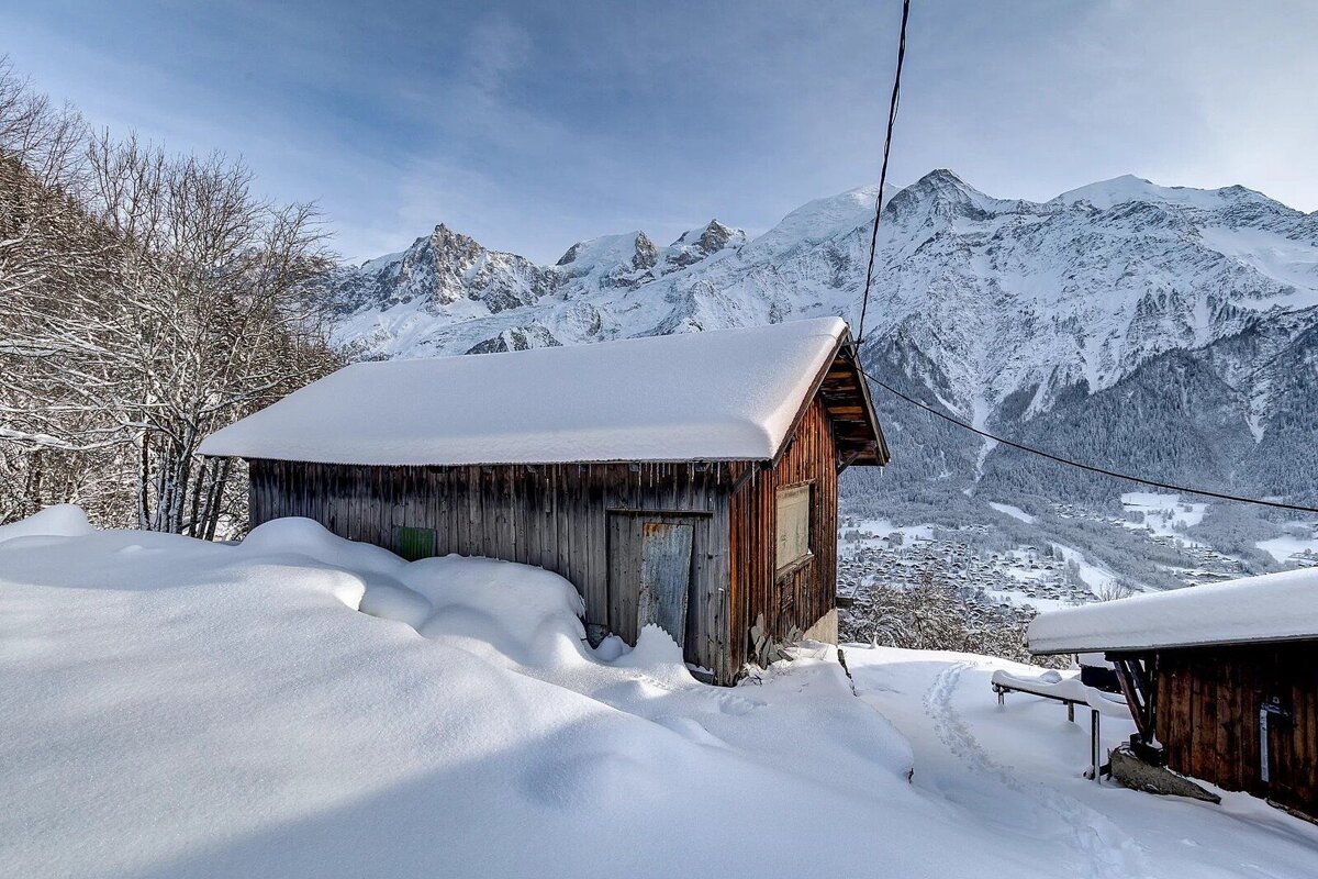 A rustic wooden cabin blanketed in deep snow, set against a stunning backdrop of towering, snow-capped mountains and a clear sky.