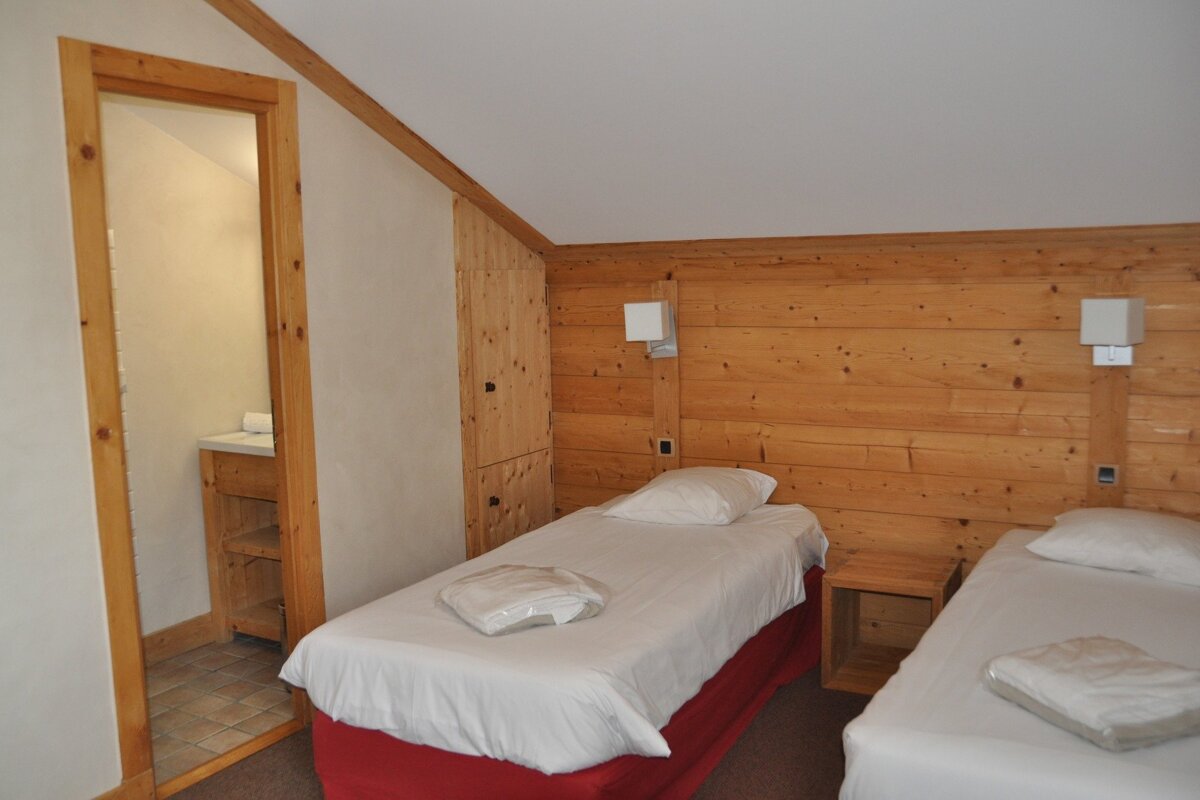 A twin bedroom features wood-paneled walls, a sloped ceiling, and two beds with white linens and red bases. A doorway reveals a vanity area.