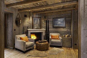 A cozy, rustic wooden room features a lit fireplace, two patterned armchairs, art, and ski lodge decor, creating a warm ambiance.