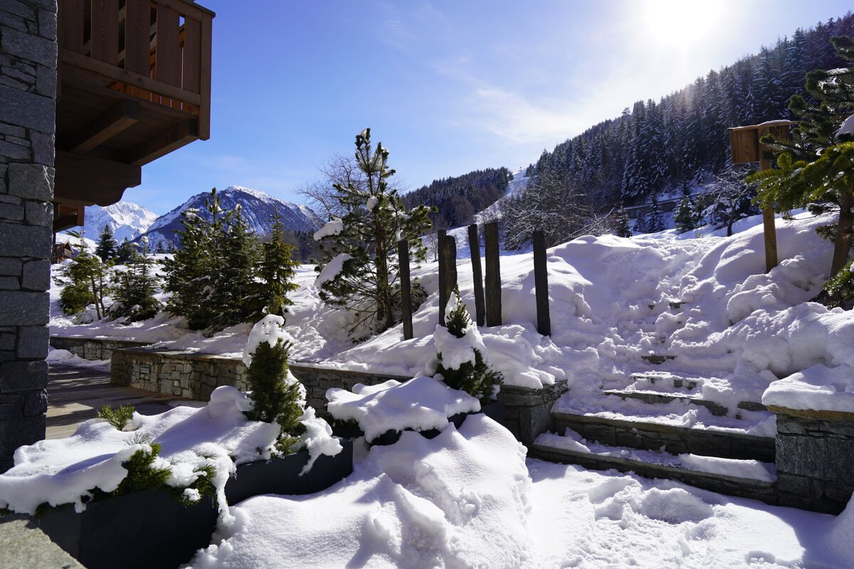 A sunny winter scene outside an alpine chalet, featuring thick snow covering stone stairs and pine trees against a mountainous backdrop.